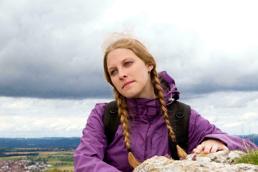 Frau mit Z&ouml;pfen in lilafarbener Jacke steht auf Berg, bew&ouml;lkter Himmel im Hintergrund. Fotografie.